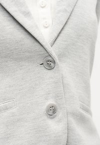 Κοντινό πλάνο γκρι υφασμάτινου σακακιού με υφή, με δύο γκρι κουμπιά κουμπωμένα πάνω από ένα λευκό πουκάμισο με κουμπιά.