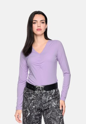 Woman with straight dark hair wearing a light purple long-sleeve top and black and white patterned pants with a black belt, standing against white background.