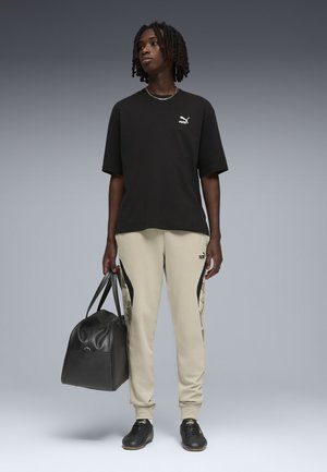 Persona con t-shirt nera Puma, pantaloni sportivi beige Puma, sneakers nere, che tiene una grande borsa da viaggio nera, in piedi davanti a uno sfondo grigio.