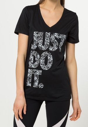 Femme portant un t-shirt noir à col en V avec un grand texte blanc texturé disant "JUST DO IT." et des leggings noirs avec des rayures diagonales blanches.