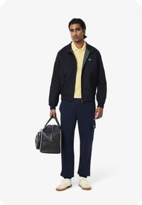 Giacca bomber blu con fodera a scacchi, polo gialla, joggers blu con tasca laterale, borsa nera e sneakers crema.