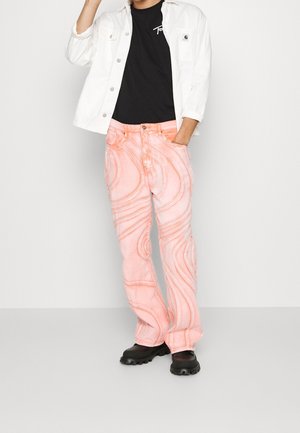 Person iført hvid jakke, sort t-shirt, lyserøde jeans med virvelmønstre, sorte klumpede sko, poserer med den ene hånd i lommen.