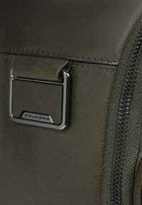 Donkergroene textuur van een tas met een zilverkleurige metalen ritssluiting en een rechthoekig metalen element met de merknaam "Piquadro".