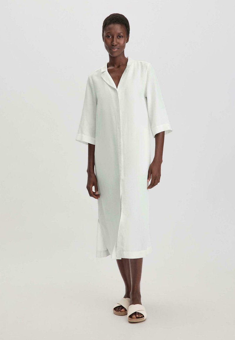 Lang lichtgroen jurkje met een V-hals, driekwart mouwen en zijslits. Gecombineerd met beige slide sandalen. Glad textuur van de stof.