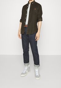 Mann mit dunkelgrüner Knopfleiste, grauem T-Shirt, dunkelblauen hochgekrempelten Jeans, braunem Gürtel und grauen High-Top-Sneakern, der vor einem weißen Hintergrund steht.