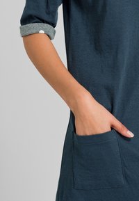 Robe turquoise avec des poignets gris retroussés, dotée d'une poche sur le côté gauche. Le tissu semble doux avec une texture lisse.