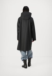 Lång svart vattentät kappa med huva, med en slät textur och en avslappnad passform, tillsammans med ljusblå baggy jeans och svarta spännbuckleskor.