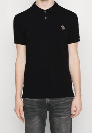 Personne portant un polo noir à manches courtes avec un petit logo zèbre multicolore sur le côté gauche de la poitrine et un jean noir délavé.