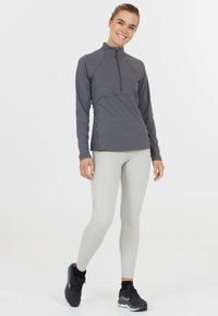 Langärmliges, mit Reißverschluss versehenes Sportoberteil in Dunkelgrau, aus glattem Stoff hergestellt. Kombiniert mit hellgrauen, eng anliegenden Leggings und schwarzen Sportschuhen.