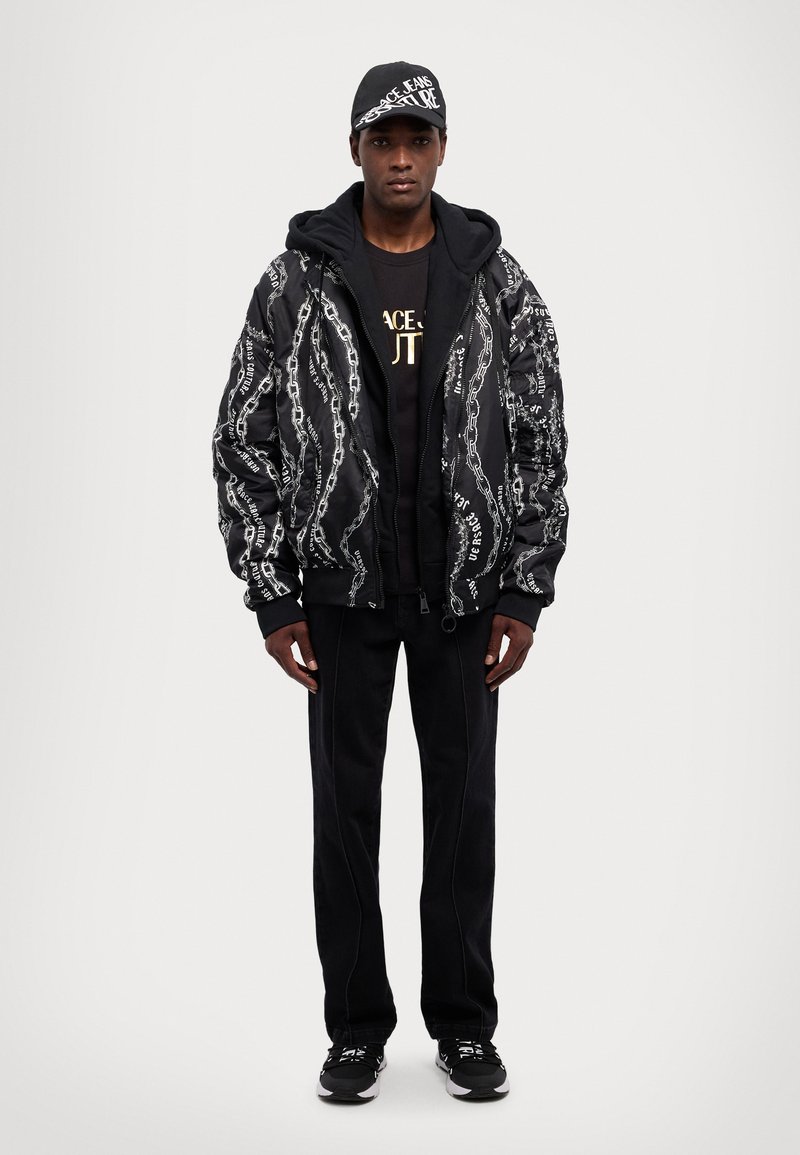 Giacca bomber nera con motivo a catena bianca, felpa nera con cappuccio, pantaloni neri e sneaker nere. Cappellino nero con dettaglio del logo bianco.