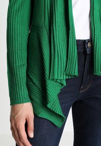 Grön ribbad kardigan med draperad design, med långa ärmar och en texturerad yta, i kombination med mörka jeans.