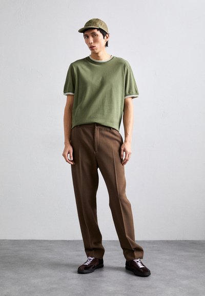 T-shirt verde oliva a maniche corte, colletto a righe bianche, abbinato a pantaloni marroni su misura. Il modello indossa un cappellino verde oliva coordinato e sneaker scure.