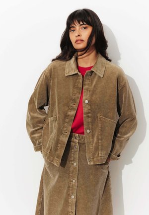 Veste en velours côtelé marron avec un design à boutons sur le devant, de grandes poches et un col, associée à une jupe assortie et un haut rouge.