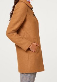 Tan uldblandingsfrakke med rund krave, to frontlommer og knaplukning, med en glat tekstur og minimalistisk design.