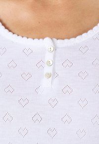 Top de punto blanco con escote festoneado, tapeta de tres botones y un estampado de corazones con pequeños recortes en toda la tela.