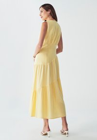 Brezrokavni rumen maksi obleki s tiered dizajnom, mehkim blagom in prilegajočim korzetom. Obleka se odlikuje z lebdečo krilo in skritimi stranskimi žepi.