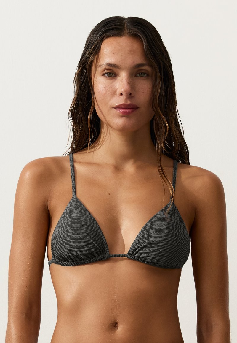 Femme aux cheveux mouillés portant un haut de bikini triangle texturé gris foncé sur un fond clair uni.