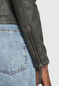 Close-up van een mouw van een zwart leren jack met ritsdetail boven een lichtblauwe spijkerbroekzak en een bruingetinte hand.