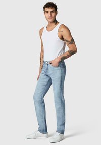 Homme avec des tatouages portant un débardeur blanc, un jean bleu clair et des baskets blanches, debout avec une main dans la poche sur fond gris.