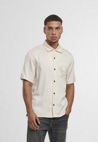 Camisa de manga corta color crema con cuello tipo botón, que presenta un bolsillo frontal con un logo contorneado en negro y botones negros.