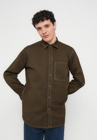 Ung man med lockigt mörkt hår, klädd i en olivgrön skjorta med knappar och bröstficka samt blå jeans, stående mot en vit bakgrund.