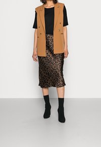 Kobieta ubrana w czarny t-shirt, camelowy brązowy bezrękawnik, midi spódnicę w panterkę oraz czarne botki na obcasie, stojąca na tle białej ściany.