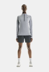 Helles graues langärmliges Sportshirt mit einem Frontreißverschluss und schwarzem Logo, kombiniert mit dunklen Shorts und weißen Sportschuhen.