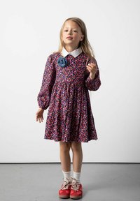 Kwiatowa sukienka z długimi rękawami i białym kołnierzykiem, z niebieskim dodatkiem w kształcie kwiatu. Czerwone buty i białe skarpetki dopełniają strój.