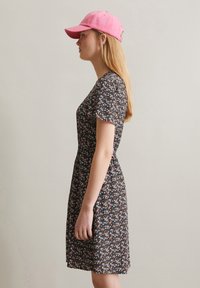 Bloemendress in donkere stof met korte mouwen, een strakke taille en kleurrijke bloemenpatronen; gecombineerd met een lichtroze baseballcap.