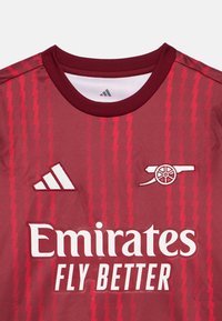 Burgundi spordisärk ümmarguse kaelusega, millel on diagonaalsed triibud, broderitud valged logod Adidaselt ja Arsenalilt ning tekst "Emirates FLY BETTER".