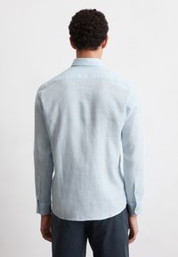 Uomo con capelli ricci che indossa una camicia a maniche lunghe blu chiaro e pantaloni scuri, rivolto di spalle su uno sfondo bianco uniforme.