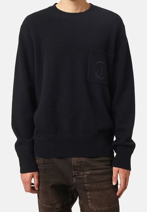 Mann trägt einen schwarzen Pullover mit langen Ärmeln und einer Brusttasche sowie dunkelbraune Hose, steht vor einem einfarbigen Hintergrund.