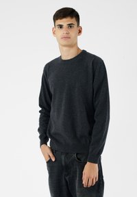 Ciemnoszara sweter dziany z okrągłym dekoltem, prążkowanymi mankietami i dołem. Model w czarnych dżinsach, z rękami w kieszeniach, na jasnym tle.