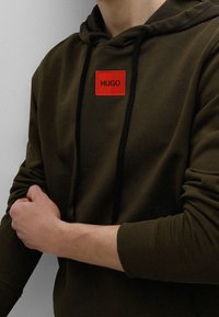 Mikina s kapucí v tmavě olivové barvě s černými šňůrkami a červenou čtvercovou nášivkou s nápisem "HUGO" na hrudi. Měkká textura.