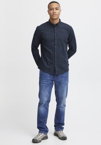 Camisa de botones azul oscuro con cuello, combinada con jeans azul claro y zapatillas grises, exhibida en un modelo de pie contra un fondo blanco.