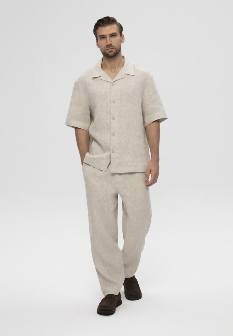 Homme marchant portant une chemise beige à manches courtes et boutons, et un pantalon ample assorti avec des chaussures brun foncé sur un fond blanc uni.