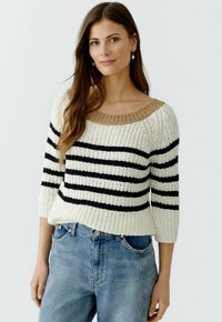 Pull en tricot crème avec des rayures noires et un col en côtes beige. Doté de manches trois-quarts et d'une coupe décontractée. Porté avec un jean bleu.