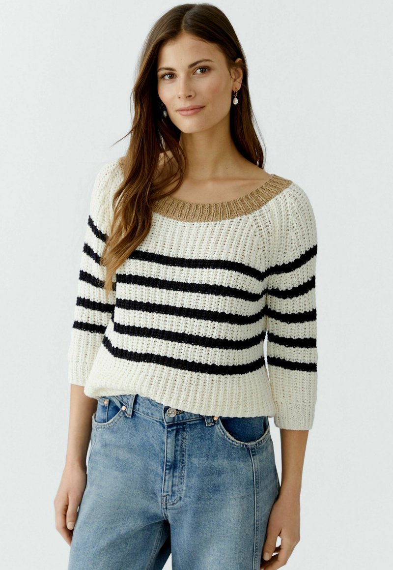 Pull en tricot crème avec des rayures noires et un col en côtes beige. Doté de manches trois-quarts et d'une coupe décontractée. Porté avec un jean bleu.