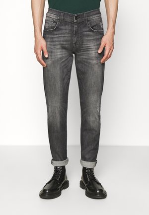 Homme portant un jean slim noir délavé avec les revers roulés et des bottes noires à lacets, debout devant un fond clair uni.