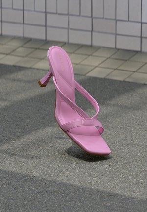 Roze sandaal met hoge hak, dunne riempjes en een vierkante neus, geplaatst op asfalt nabij een betegelde muur als achtergrond.