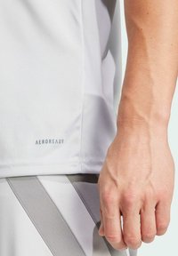 Lichtgrijs sportshirt met AEROREADY-opdruk aan de onderkant. De stof is glad, ontworpen voor prestaties, met een close-up van een hand.