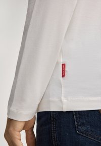 Nahaufnahme einer Hand einer Person, die ein weißes langärmeliges Hemd mit einem roten JOOP! JEANS-Label trägt, kombiniert mit dunkelblauen Jeans.