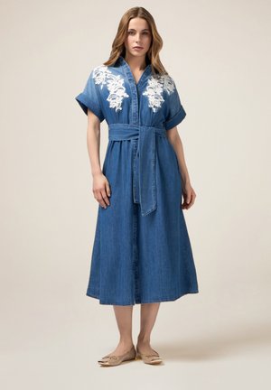 Vestido de mezclilla con mangas cortas, cintura ceñida con cinturón y bordado floral blanco en el pecho. Silueta larga y fluida. Color azul claro.