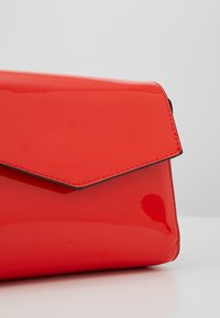 Bolso de mano de charol rojo con cierre de solapa y costuras en contraste negras. Presenta una textura lisa y brillante y un diseño anguloso.