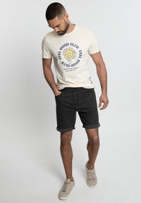 T-shirt beige avec un motif de soleil et un texte bleu, associé à un short en denim noir et des baskets grises. Look décontracté, ourlet roulé sur le short.
