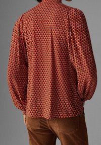 Rode blouse met lange mouwen en een geometrisch patroon met zwarte en crèmewitte accenten. Zachte stof, ruime pasvorm en geplooide schouders.