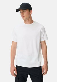 Hvid bomulds t-shirt med korte ærmer, rund hals og et diskret logo på brystet. Kombineret med en marineblå baseball cap.