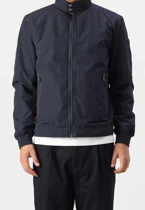 Mand iført en marineblå jakke med lynlås og høj krave, lynlåslommer i siden og ribstrikkede ærmekanter, over en hvid skjorte og sorte bukser.