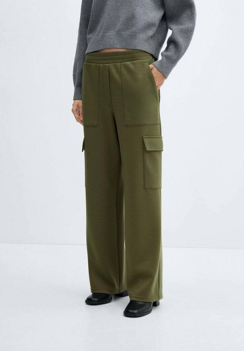 Mujer vestida con pantalones cargo de pierna ancha en verde oliva, con bolsillos frontales y laterales, suéter gris corto y botas negras, de pie contra un fondo blanco.