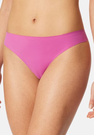 Frau trägt nahtlose, rosafarbene Bikini-Unterwäsche und zeigt die Körpermitte sowie die oberen Oberschenkel vor weißem Hintergrund.